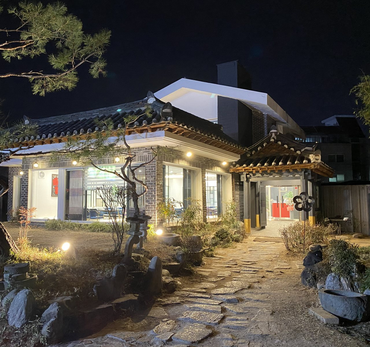 밤 정원과 한옥 외관 — 조명 켠 소나무·석등·돌길, 기와지붕 아래 흰 외벽과 통유리
